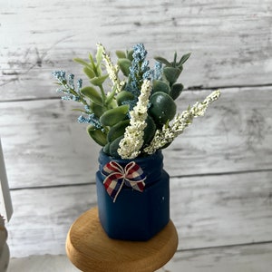 Pode incluir: Um pequeno frasco azul com um la&ccedil;o vermelho, branco e azul, cheio de um buqu&ecirc; de flores artificiais brancas e azuis. O frasco est&aacute; sentado em um pedestal de madeira.