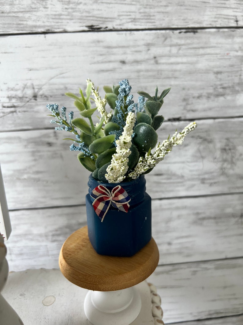 Pode incluir: Um pequeno frasco azul com um la&ccedil;o vermelho, branco e azul, cheio de um buqu&ecirc; de flores artificiais brancas e azuis. O frasco est&aacute; sentado em um pedestal de madeira.