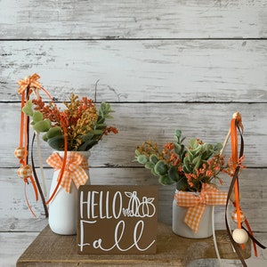 May include: Two white ceramic vases with orange and white gingham ribbon bows, filled with fall foliage, sit on a wooden shelf with a brown wooden sign that reads "Hello Fall" with a white pumpkin graphic.