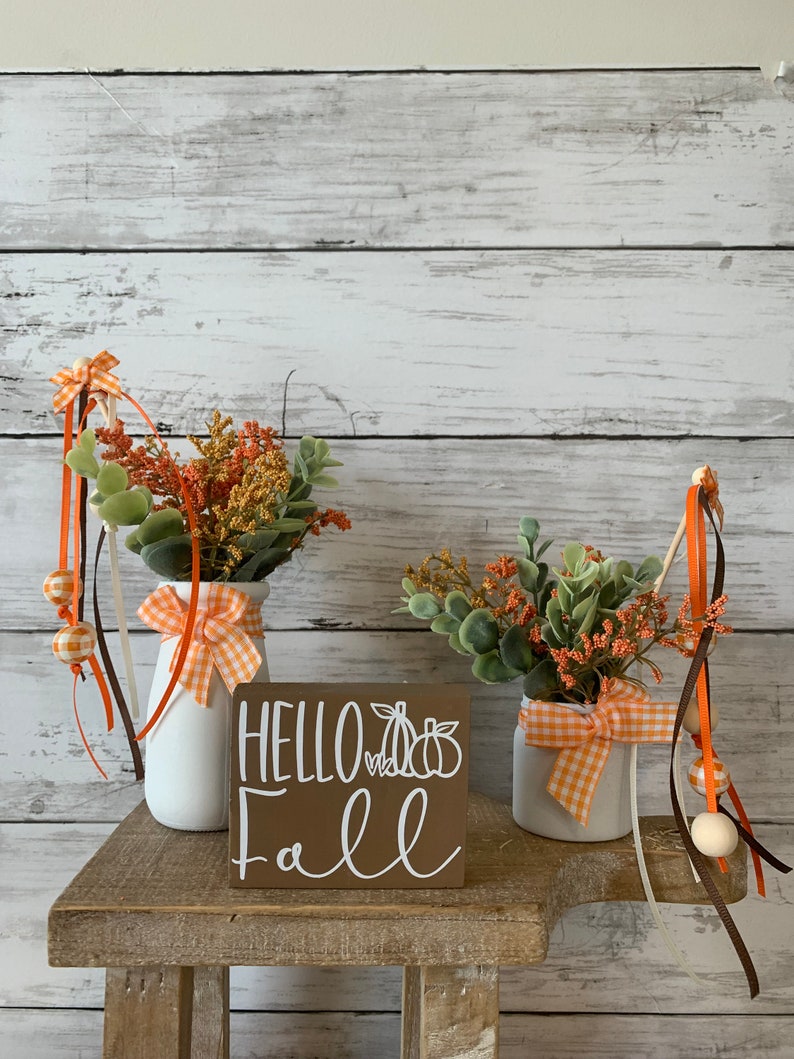 May include: Two white ceramic vases with orange and white gingham ribbon bows, filled with fall foliage, sit on a wooden shelf with a brown wooden sign that reads "Hello Fall" with a white pumpkin graphic.