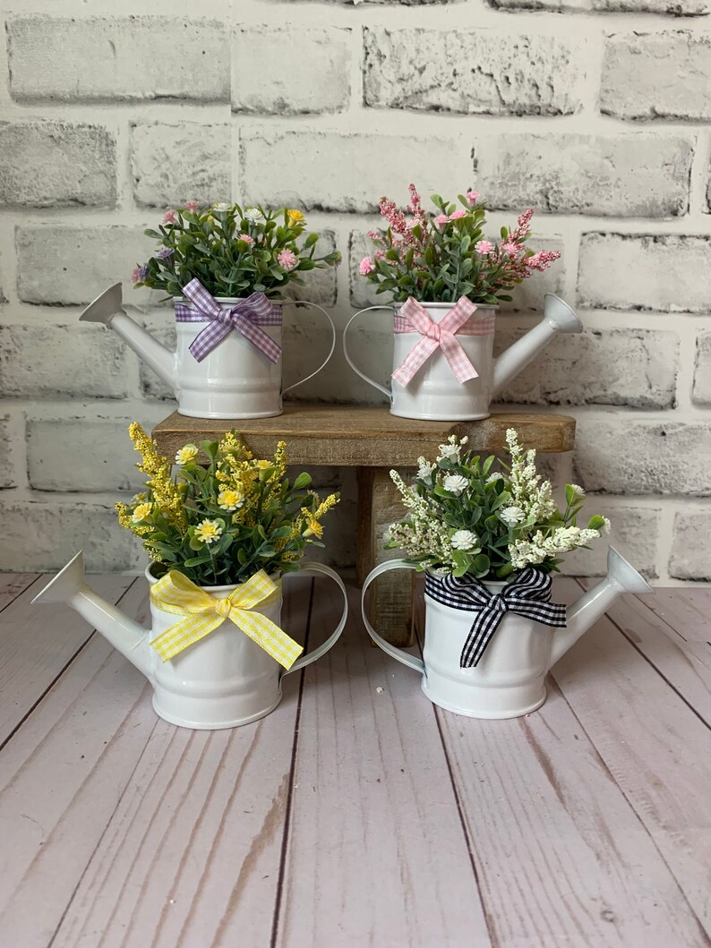 May include: Four white metal watering cans filled with colorful faux flowers. Each can has a different colored gingham bow: purple, pink, yellow, and black. The watering cans are arranged on a wooden surface against a white brick wall.