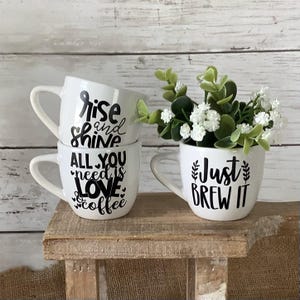 May include: Three white ceramic mugs with black lettering. The top mug says "Rise and Shine", the middle mug says "All You Need is Love & Coffee", and the bottom mug says "Just Brew It".