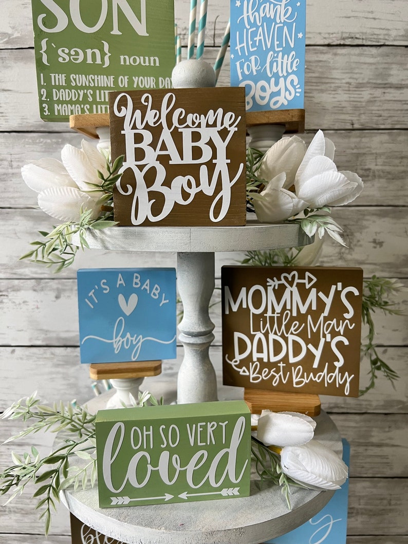 K&ouml;nnte beinhalten: Ein dreistufiges Tablett, das mit Holzschildchen f&uuml;r eine Babyparty dekoriert ist. Die Schilder zeigen S&auml;tze wie "Willkommen Baby Boy", "Es ist ein Baby Boy", "Mamas kleiner Mann Papas bester Freund" und "So sehr geliebt". Das Tablett ist mit wei&szlig;en Blumen und Gr&uuml;npflanzen dekoriert.