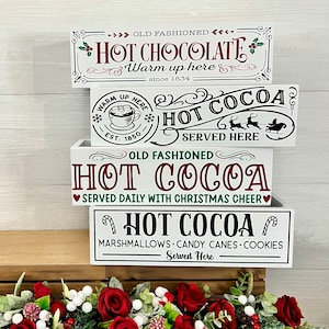 May include: Three white wooden boxes stacked on top of each other. Each box has a different design with the words "Hot Cocoa" and Christmas-themed graphics. The top box says "Old Fashioned Hot Chocolate" and "Warm up here since 1834". The middle box says "Hot Cocoa Served Here" with a Christmas sleigh and reindeer. The bottom box says "Old Fashioned Hot Cocoa Served Daily with Christmas Cheer" and "Hot Cocoa Marshmallows Candy Canes Cookies Served Here".