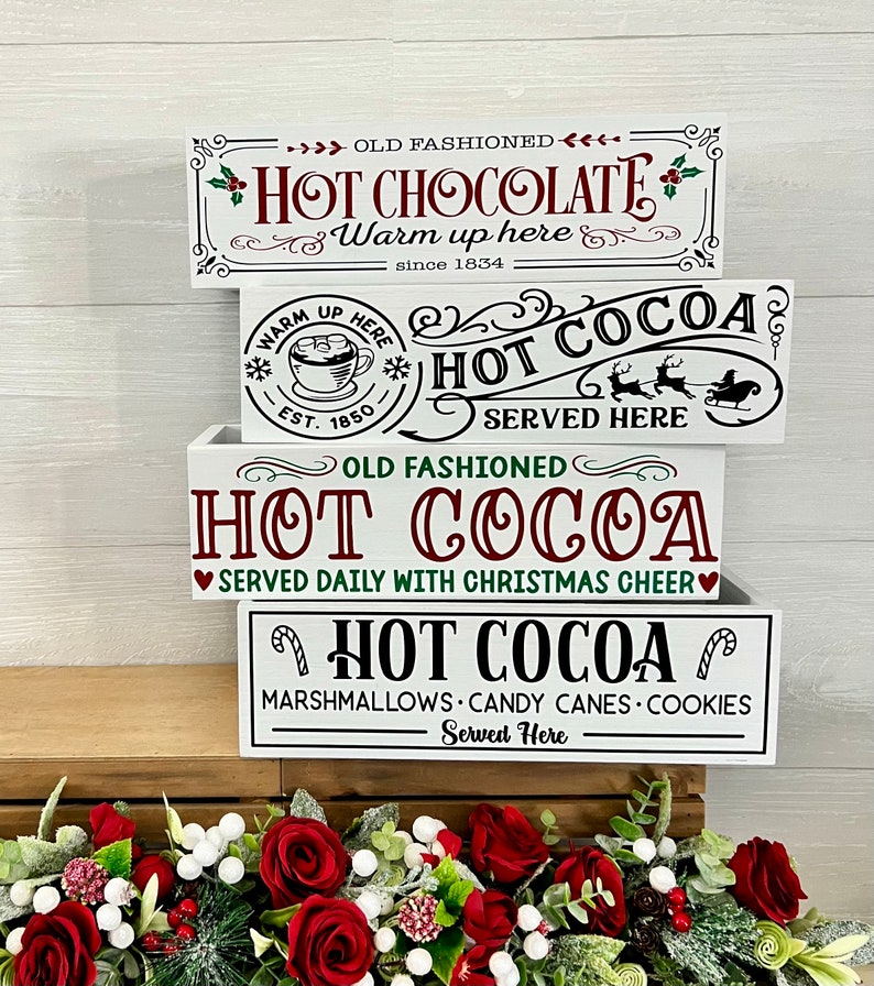 May include: Three white wooden boxes stacked on top of each other. Each box has a different design with the words "Hot Cocoa" and Christmas-themed graphics. The top box says "Old Fashioned Hot Chocolate" and "Warm up here since 1834". The middle box says "Hot Cocoa Served Here" with a Christmas sleigh and reindeer. The bottom box says "Old Fashioned Hot Cocoa Served Daily with Christmas Cheer" and "Hot Cocoa Marshmallows Candy Canes Cookies Served Here".
