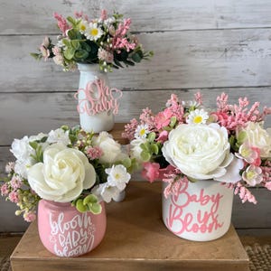 May include: Three pink and white floral arrangements in different sized jars. The jars are decorated with pink lettering. The first jar says "Oh Baby", the second jar says "Bloom Baby Bloom", and the third jar says "Baby Be in Bloom".