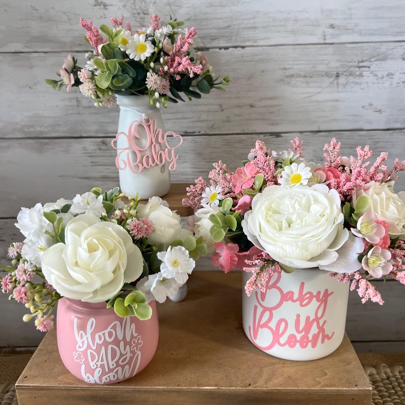 May include: Three pink and white floral arrangements in different sized jars. The jars are decorated with pink lettering. The first jar says "Oh Baby", the second jar says "Bloom Baby Bloom", and the third jar says "Baby Be in Bloom".