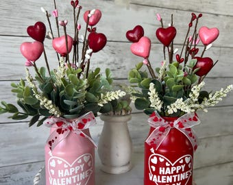 May include: Two small glass jars decorated with hearts and the text "Happy Valentine's Day" are filled with faux greenery and pink and red heart-shaped decorations. The jars are sitting on a white tiered stand.