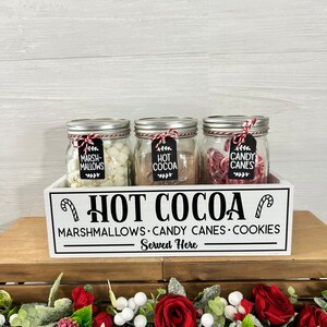 May include: White wooden box with the text "HOT COCOA" and "MARSHMALLOWS CANDY CANES COOKIES Served Here". Three glass jars with lids are inside the box. The jars are filled with marshmallows, chocolate, and candy canes. The jars have labels that say "Marshmallows", "Hot Cocoa", and "Candy Canes".