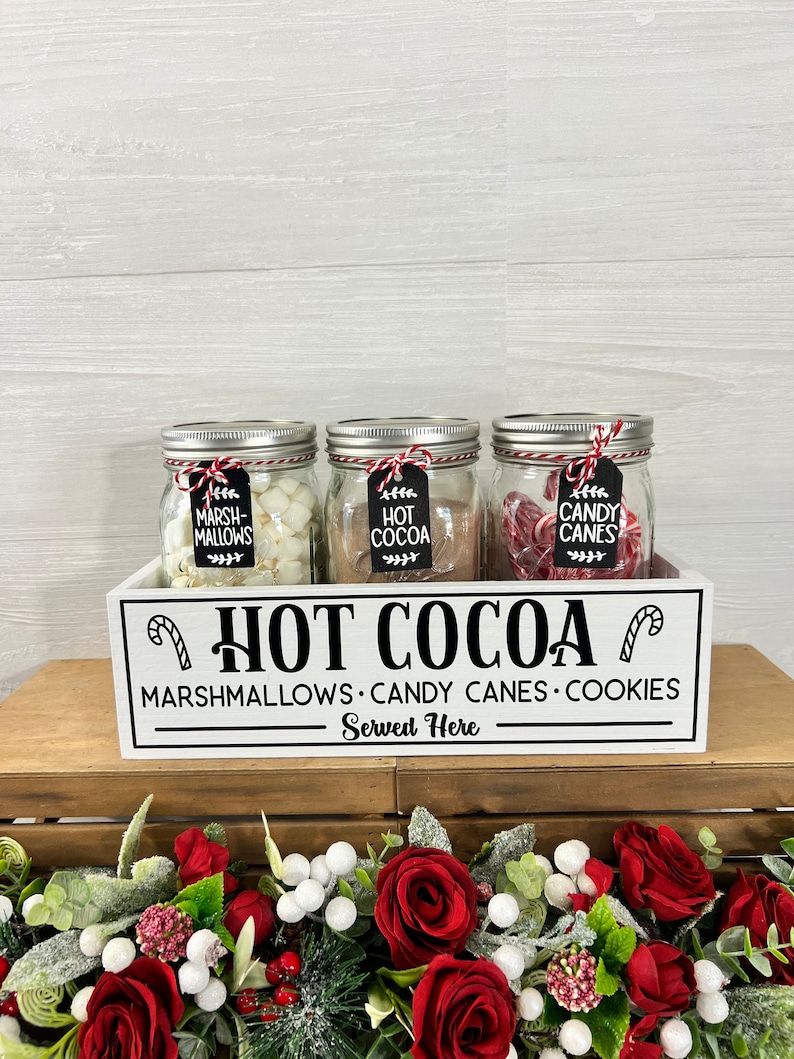 May include: White wooden box with the text "HOT COCOA" and "MARSHMALLOWS CANDY CANES COOKIES Served Here". Three glass jars with lids are inside the box. The jars are filled with marshmallows, chocolate, and candy canes. The jars have labels that say "Marshmallows", "Hot Cocoa", and "Candy Canes".