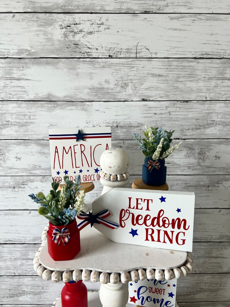 Pode incluir: Uma bandeja de tr&ecirc;s n&iacute;veis decorada com decora&ccedil;&atilde;o patri&oacute;tica vermelha, branca e azul. O n&iacute;vel superior apresenta uma placa que diz "Let freedom ring" com estrelas. O n&iacute;vel intermedi&aacute;rio apresenta uma placa que diz "America God Bless Grace on Us" e uma pequena planta em vaso. O n&iacute;vel inferior apresenta uma placa que diz "Home Sweet Home" e uma garrafa vermelha.