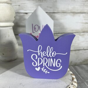 K&ouml;nnte beinhalten: Lilafarbener, tulpenf&ouml;rmiger Holzschild mit dem Text "hello spring" in wei&szlig;en Buchstaben mit einem kleinen Herz und einem Blattmotiv.