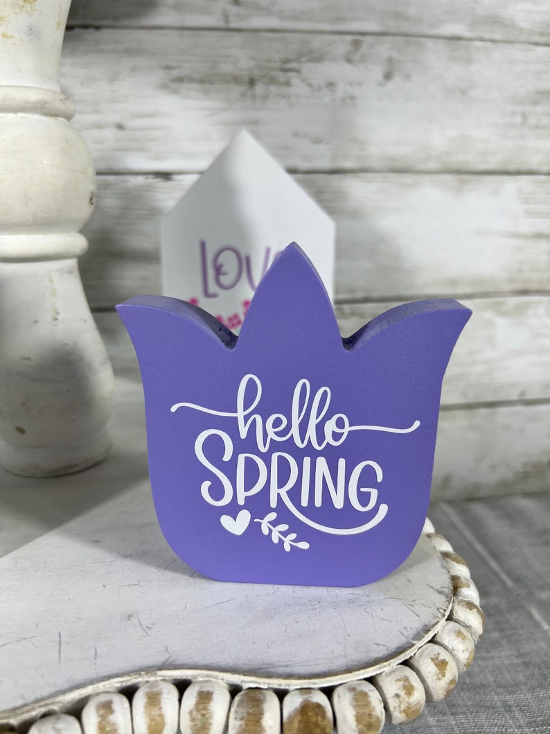 K&ouml;nnte beinhalten: Lilafarbener, tulpenf&ouml;rmiger Holzschild mit dem Text "hello spring" in wei&szlig;en Buchstaben mit einem kleinen Herz und einem Blattmotiv.