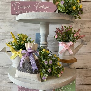 May include: A three-tiered decorative stand with floral arrangements and signs. The top tier features a sign that reads "Flower Farm" and a white watering can. The middle tier has a lavender sachet. The bottom tier has a sign that reads "Spring".