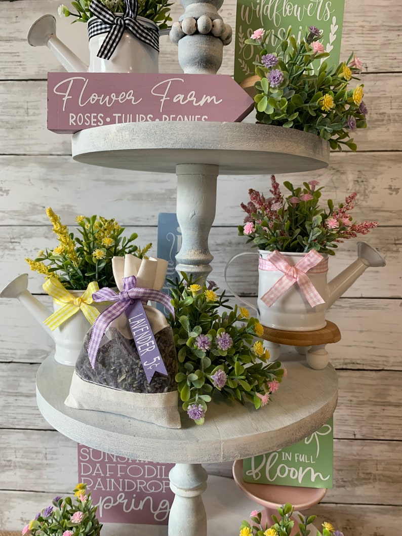 May include: A three-tiered decorative stand with floral arrangements and signs. The top tier features a sign that reads "Flower Farm" and a white watering can. The middle tier has a lavender sachet. The bottom tier has a sign that reads "Spring".