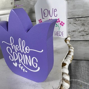 K&ouml;nnte beinhalten: Lilafarbener Tulpen-f&ouml;rmiger Holzschild mit dem Text "hello spring" in wei&szlig;en Buchstaben. Ein kleineres wei&szlig;es Holzschild mit dem Text "love here" und rosa Blumen befindet sich hinter dem Tulpenschild.