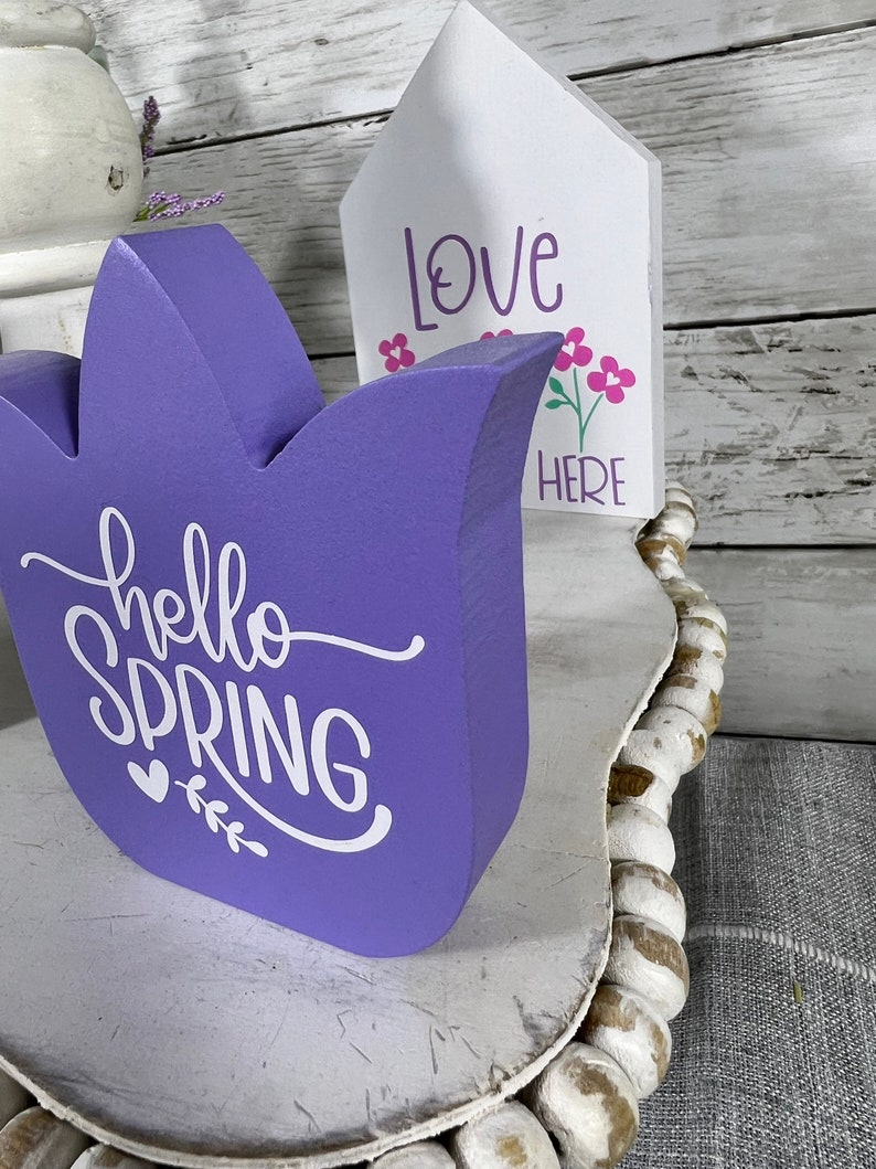 K&ouml;nnte beinhalten: Lilafarbener Tulpen-f&ouml;rmiger Holzschild mit dem Text "hello spring" in wei&szlig;en Buchstaben. Ein kleineres wei&szlig;es Holzschild mit dem Text "love here" und rosa Blumen befindet sich hinter dem Tulpenschild.