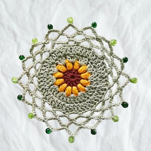 May include: A handmade, crocheted doily with a sunflower design at its centre. The sunflower has yellow petals and a maroon centre. The doily is light green with a decorative outer edge, embellished with green beads. The craftwork is set against a white background.
