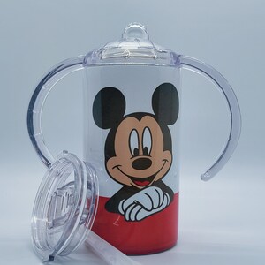 May include: A white and red sippy cup featuring Mickey Mouse. The cup has a clear lid, handles, and a straw. The design includes a smiling Mickey Mouse face on the front.