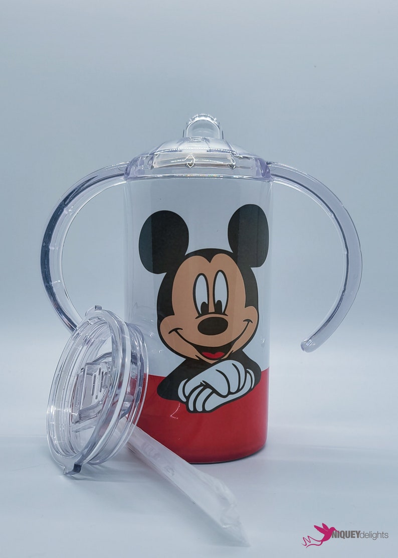 May include: A white and red sippy cup featuring Mickey Mouse. The cup has a clear lid, handles, and a straw. The design includes a smiling Mickey Mouse face on the front.