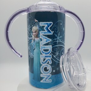 K&ouml;nnte beinhalten: Ein blau-wei&szlig;er Trinkbecher mit der Figur Elsa aus dem Film Frozen, mit dem Namen "MADISON" in Wei&szlig;. Der Becher hat lila Griffe, einen transparenten Deckel und Strohhalm. Der Becher ist mit Schneeflocken verziert.