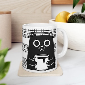 Puede incluir: Taza de cerámica blanca con una ilustración en blanco y negro de un gato sosteniendo una taza de café. El gato tiene ojos grandes y una expresión linda. La taza tiene un patrón geométrico en blanco y negro en la parte posterior.