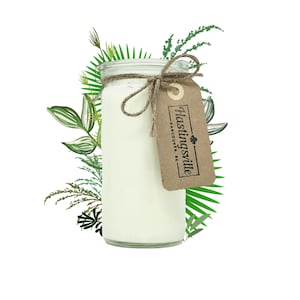 May include: A white candle in a clear glass jar with a brown paper tag that says "Hastingsville, Vancouver, BC". The jar is tied with twine and surrounded by green foliage.
