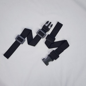 Może przedstawiać: Dwa czarne, regulowane paski z plastikowymi klamrami i suwakami. Paski wykonane są z tkanego materiału i są przeznaczone do regulacji. Klamry i suwaki są z czarnego plastiku. Paski leżą na białej powierzchni.