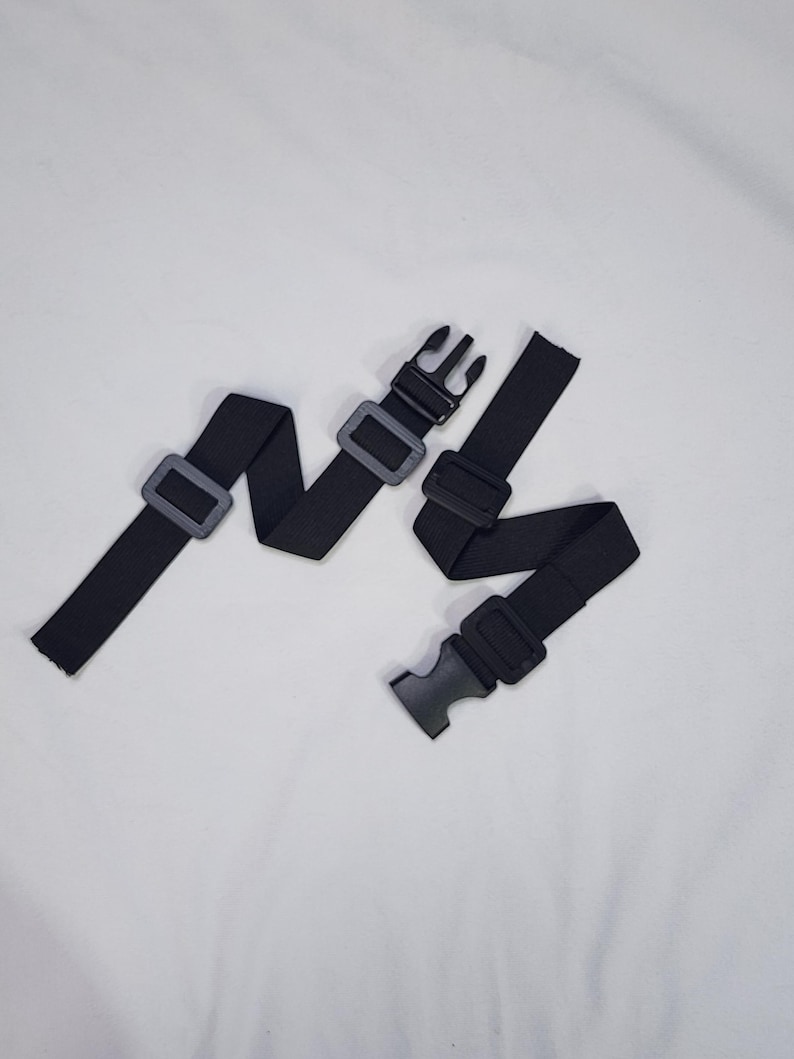 May include: Two black adjustable straps with plastic buckles and sliders. The straps are made of a woven material and are designed to be adjustable. The buckles and sliders are black plastic. The straps are laid out on a white surface.