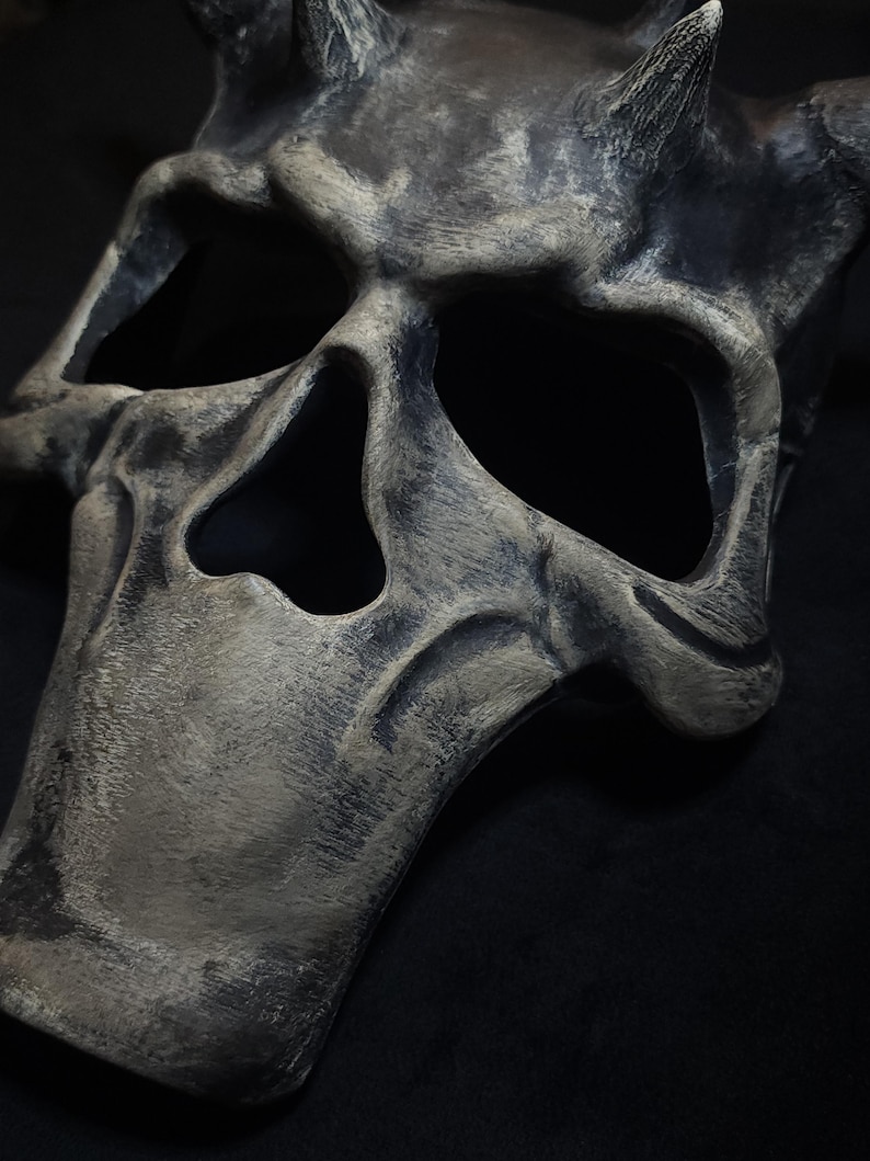 May include: A gray and black skull mask with horns. The mask has a weathered, distressed look.