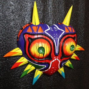 May include: A colorful, wooden mask with a heart shape and sharp, pointed edges. The mask is painted in shades of red, yellow, blue, green, and black. It has two large, round eyes with yellow irises and green pupils.