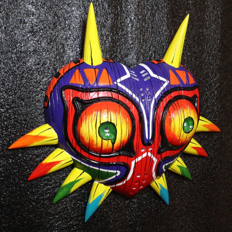 May include: A colorful, wooden mask with a heart shape and sharp, pointed edges. The mask is painted in shades of red, yellow, blue, green, and black. It has two large, round eyes with yellow irises and green pupils.
