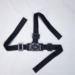 May include: A black chest harness with adjustable straps and multiple buckles. The harness is designed to secure a camera or other device, featuring a central mounting point and side adjustment buckles. The straps are made of a durable, woven material.