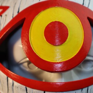 May include: A close-up of a red and yellow eye with a white background. The eye is made of a textured material and has a 3D effect.