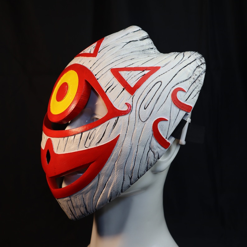 May include: A white and red mask with a wood grain texture. The mask has a large red eye with a yellow circle in the center. The mask has a red mouth and a red triangle on the forehead.