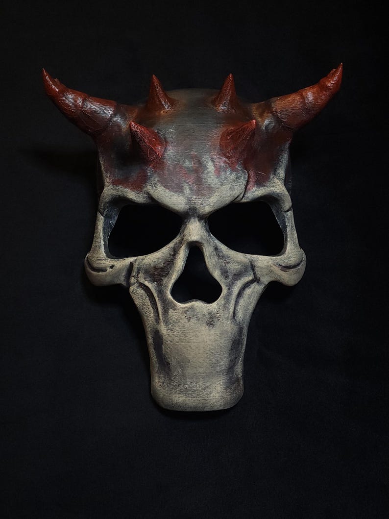 May include: A white and red skull mask with horns and spikes. The mask is made of a textured material and has a distressed look.