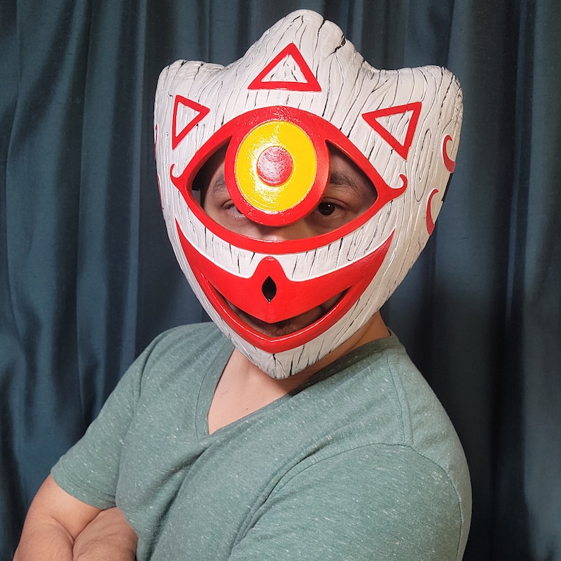 May include: A white mask with a red mouth and a large yellow eye. The mask has a wood grain texture and geometric patterns.
