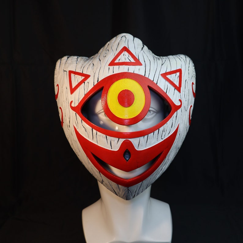 May include: A white mask with a red and yellow eye design. The mask has a wooden texture and a wide, toothy grin.
