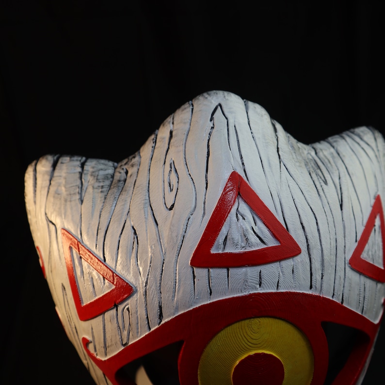 May include: A white mask with red and yellow details. The mask has a wood grain texture and a triangular pattern.