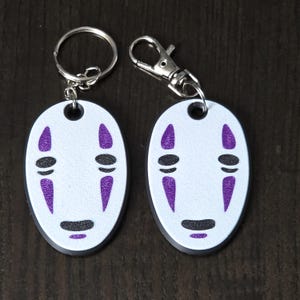 Op de afbeelding: Twee witte ovale sleutelhangers met een zwarte rand en een paars en zwart No Face-personage uit de film Spirited Away. De sleutelhangers zijn bevestigd aan zilveren sleutelringen.