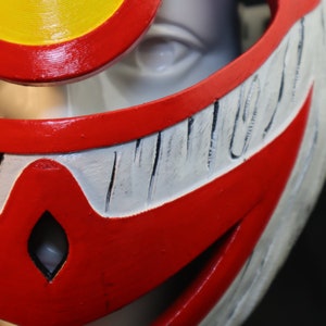 May include: Close-up of a red and white plastic mask with a yellow and red circle on top. The mask has a white, textured, and slightly raised design around the eyes and mouth.