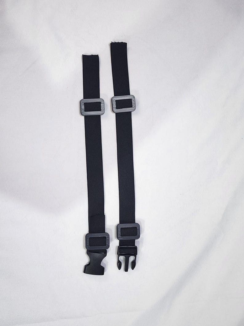 May include: Two black adjustable straps with gray plastic buckles and a black plastic clip. The straps are made of a woven material and are designed to be used with a variety of items.