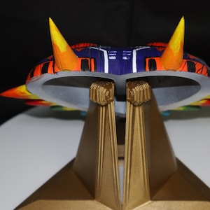 May include: A colorful, multi-layered, 3D printed model of a spaceship with a purple and black body, orange and yellow accents, and a gold stand.