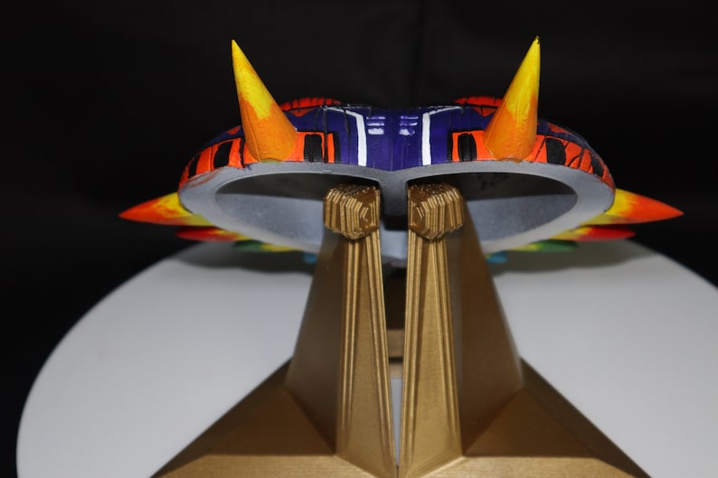 May include: A colorful, multi-layered, 3D printed model of a spaceship with a purple and black body, orange and yellow accents, and a gold stand.