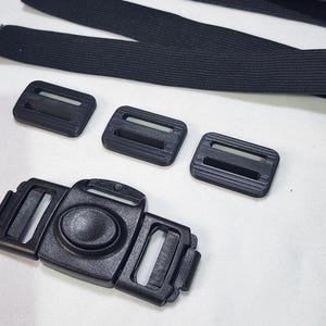 May include: Close-up of black plastic buckles, slides, and elastic straps. The image shows a variety of components, including a large center buckle with a push-in clasp, three smaller slides, and several lengths of black elastic.