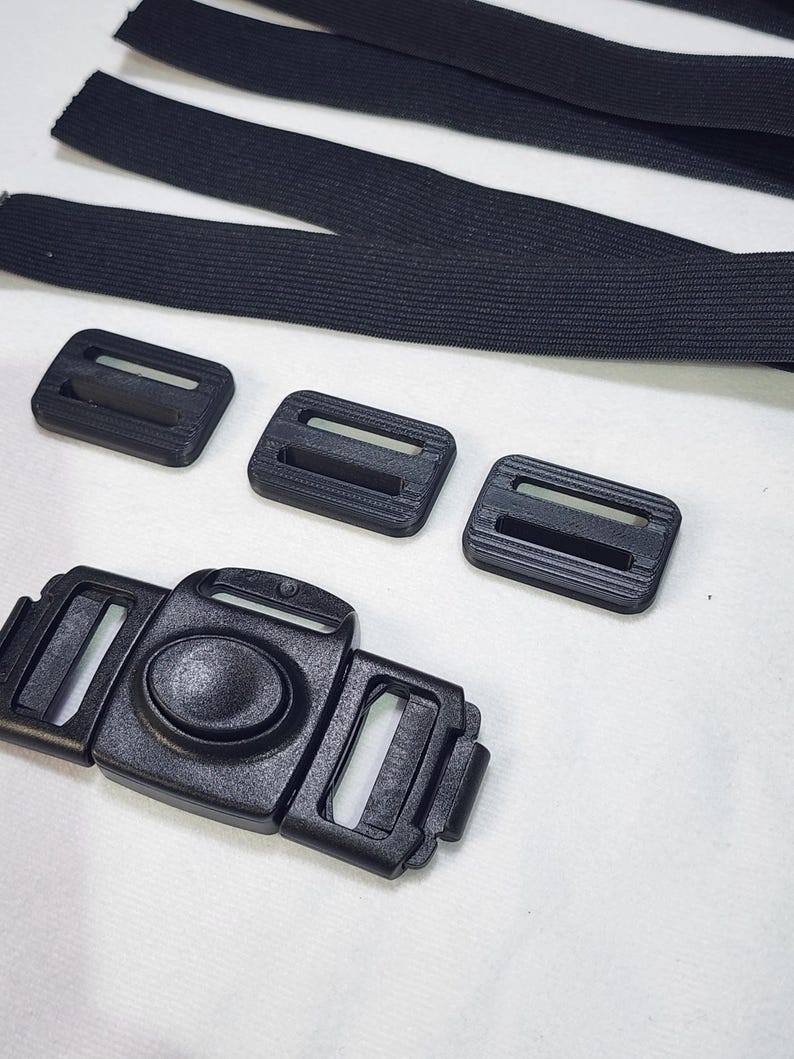 May include: Close-up of black plastic buckles, slides, and elastic straps. The image shows a variety of components, including a large center buckle with a push-in clasp, three smaller slides, and several lengths of black elastic.