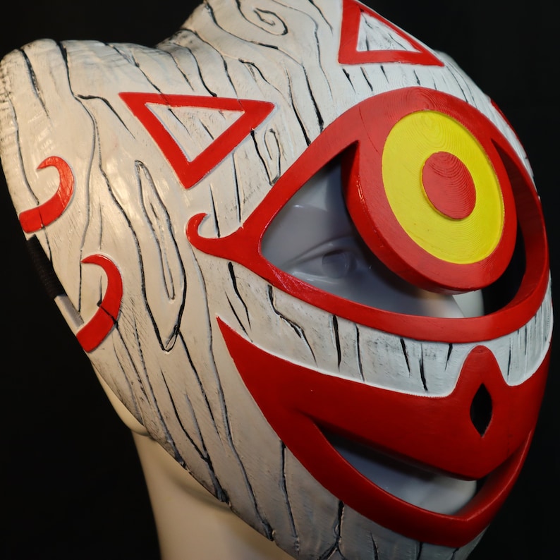 May include: A white wooden mask with red and yellow paint. The mask has a large eye with a yellow circle and a red center. The mask also has a large red smile.