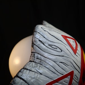May include: Close-up of a white wooden mask with red and yellow accents. The mask has a textured surface that resembles wood grain.