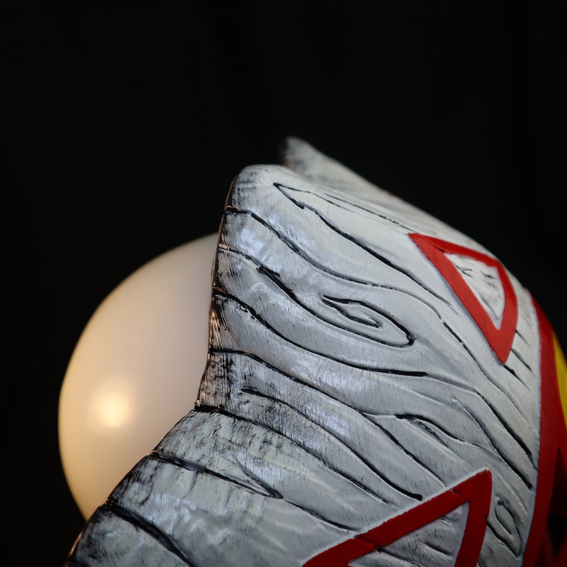 May include: Close-up of a white wooden mask with red and yellow accents. The mask has a textured surface that resembles wood grain.