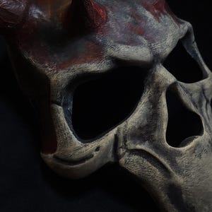 May include: A detailed, hand-painted skull mask with a textured surface and sharp, red horns. The mask is made of a hard material and has a realistic look.